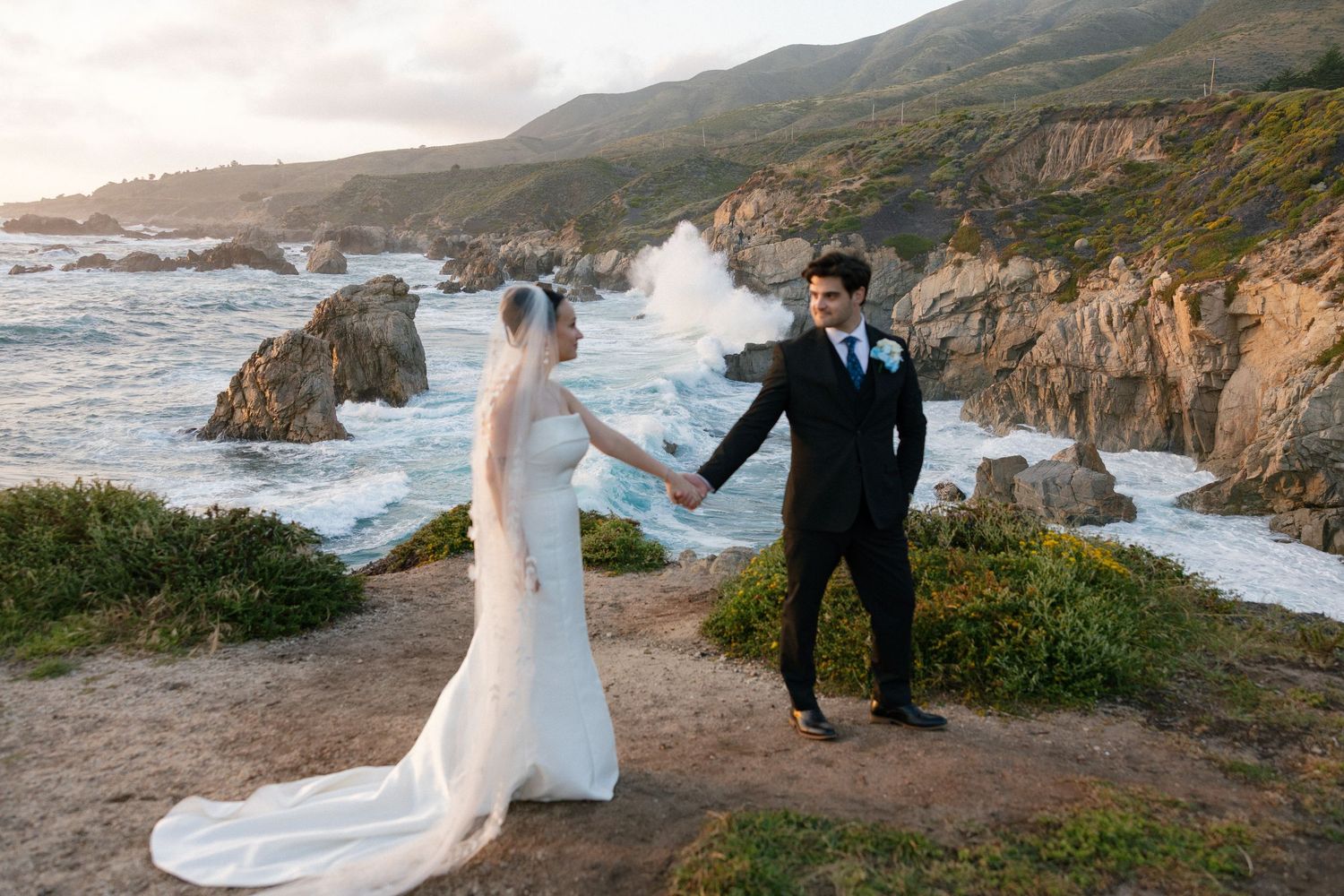 Bride and groom hold hands on rocky Big Sur coastline cliff with dramatic ocean waves and golden sunset behind them.