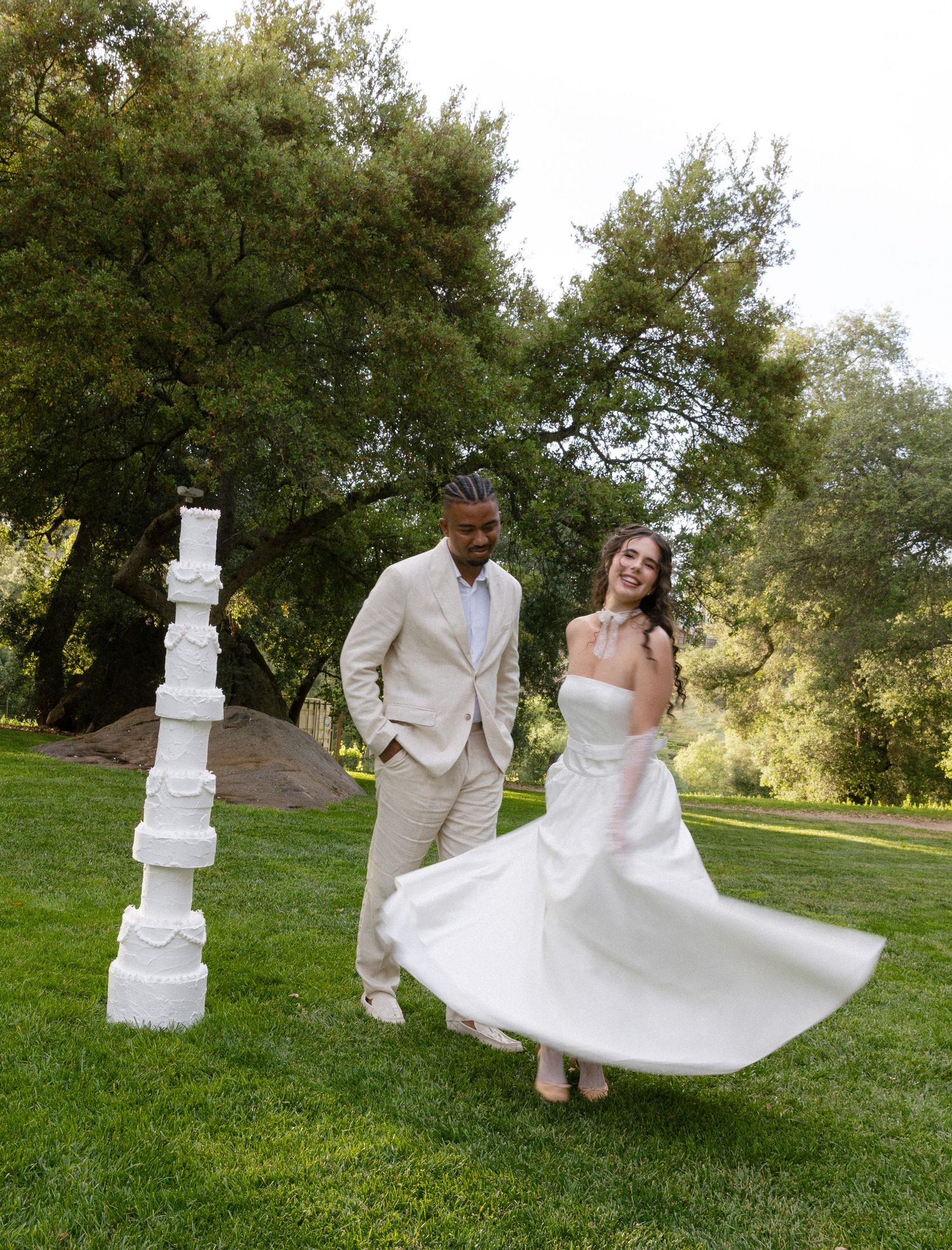 A wedding couple poses playfully near a tall white wedding cake in an outdoor garden setting during golden hour.