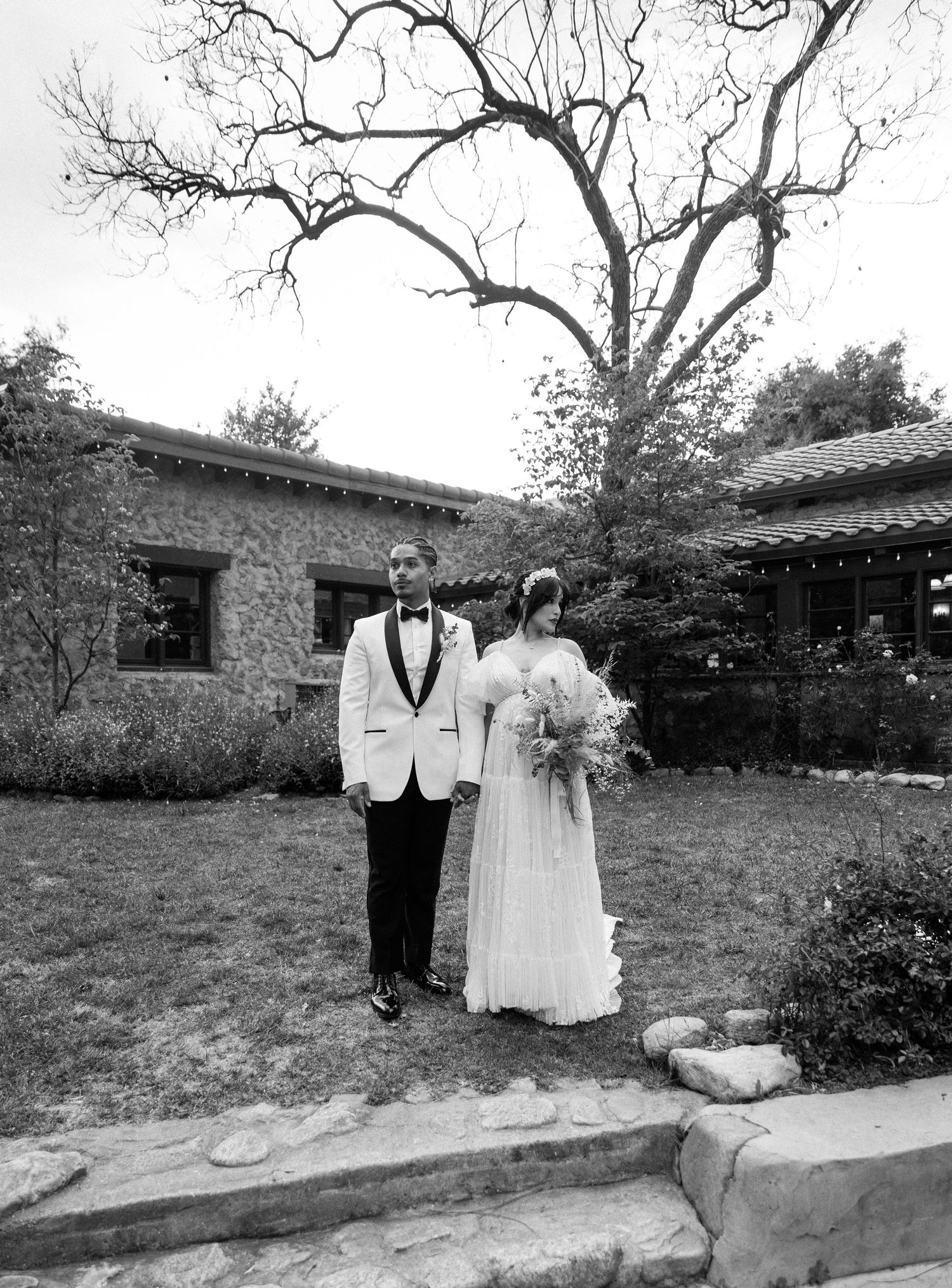 A couple in formal wedding attire poses together in a garden setting, creating a romantic black and white portrait.