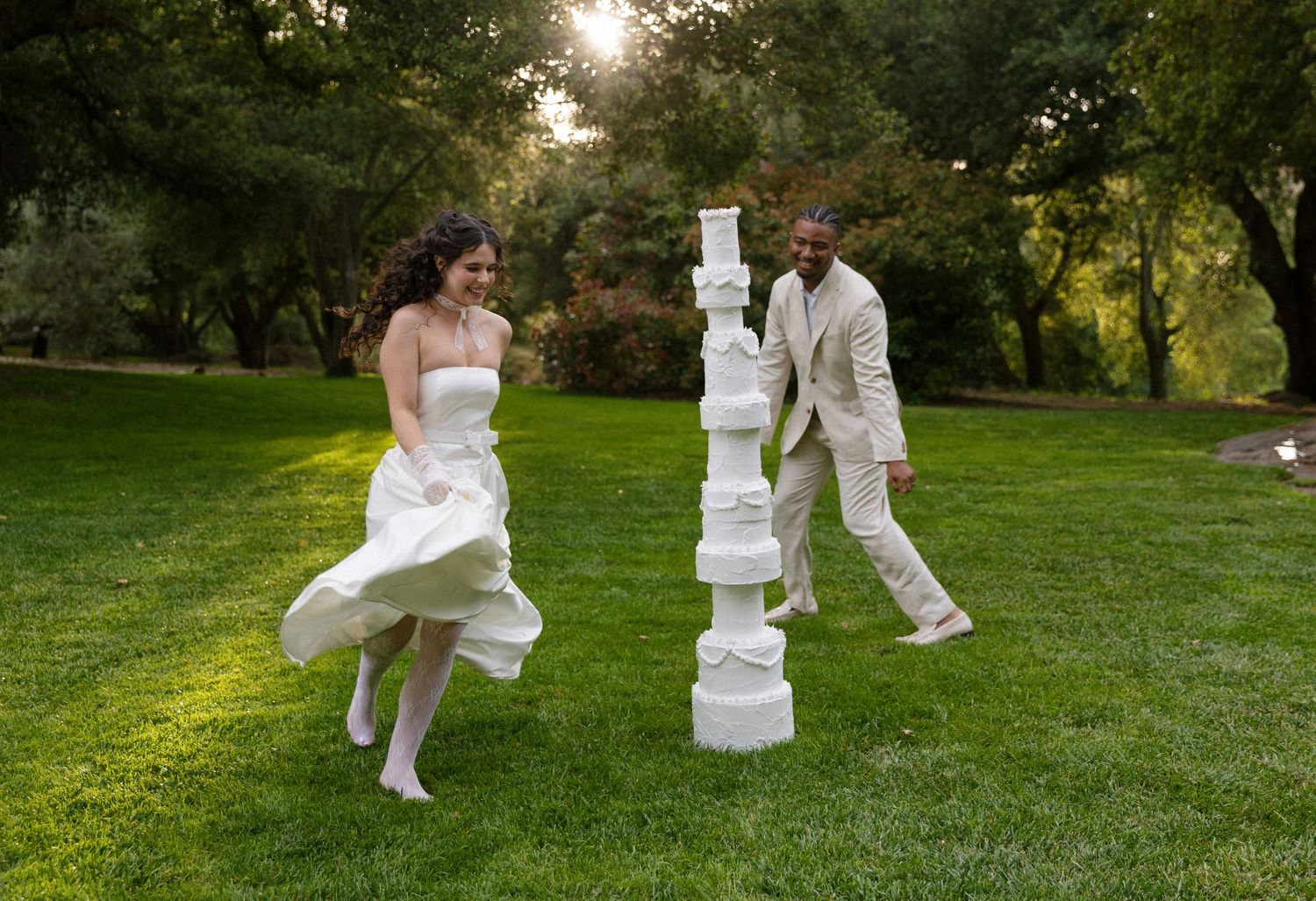 A couple in white attire plays Jenga on a grassy lawn at sunset while their clothing flows in the breeze.
