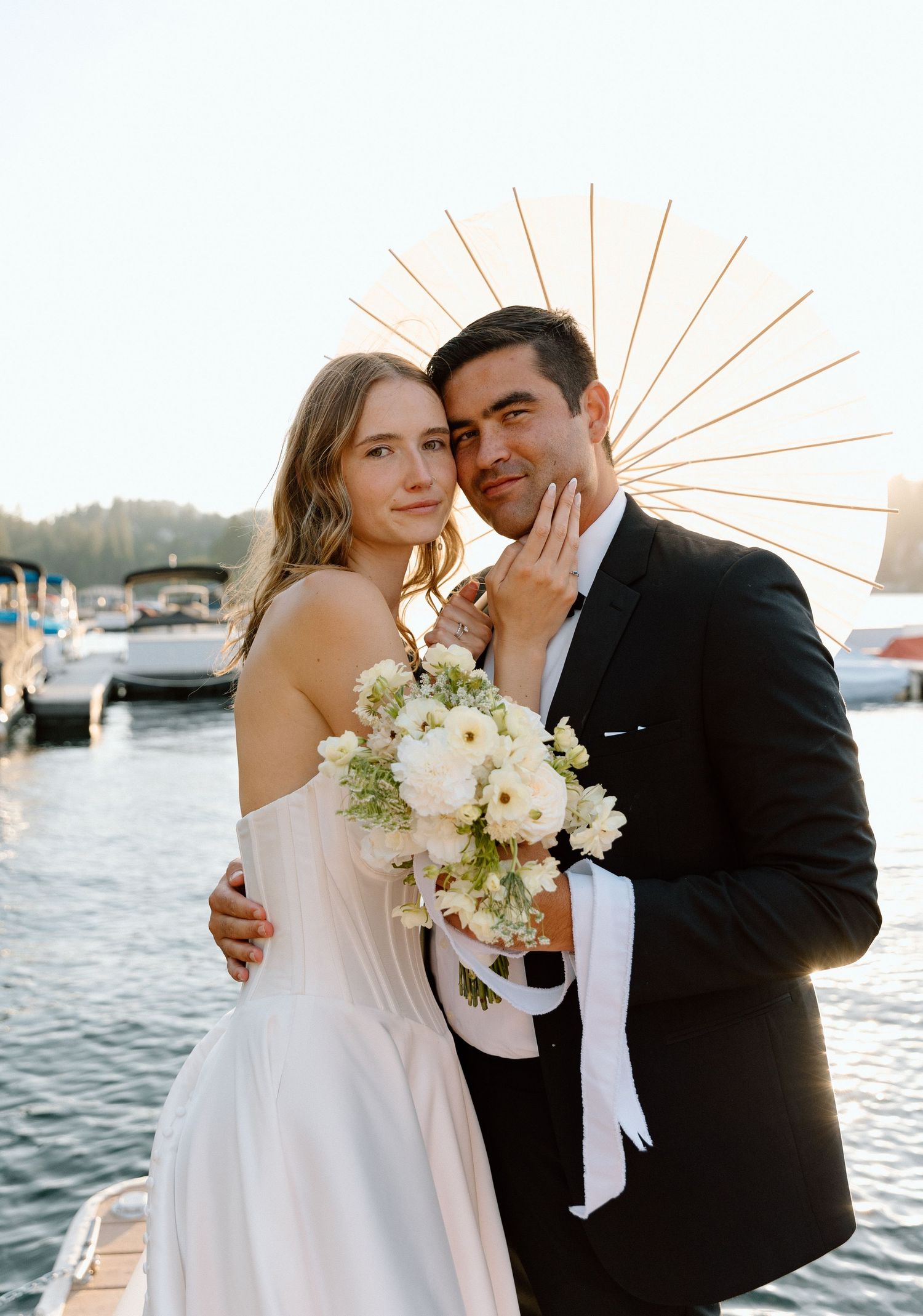 A romantic wedding photo of a couple posing by a marina at sunset with a white bridal bouquet.