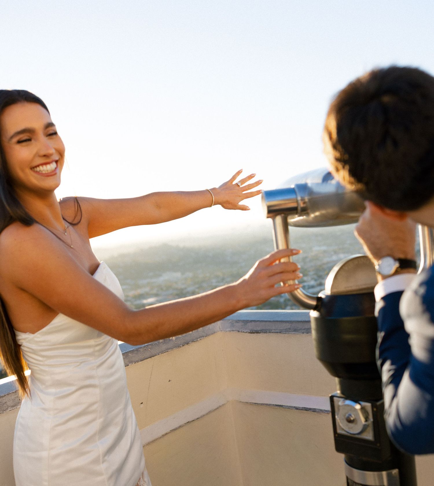 A smiling woman in a white dress gestures near a coin-operated telescope on a scenic overlook at sunset.