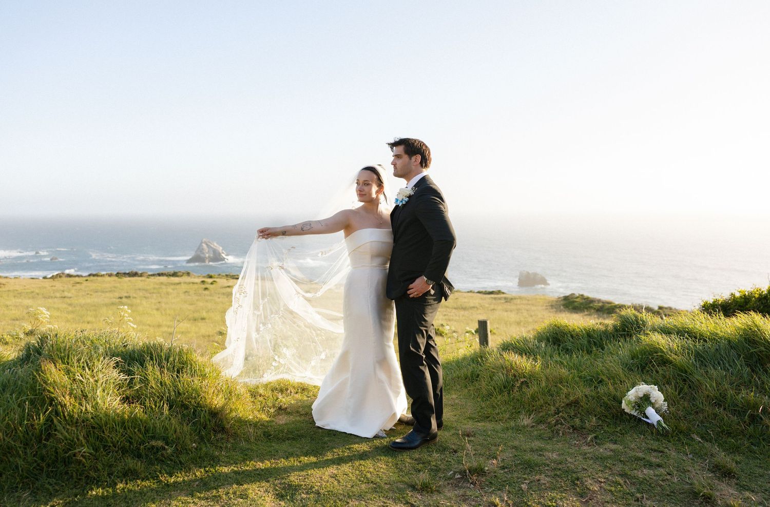 Bride and groom pose on coastal cliffside, wedding veil flowing in breeze with ocean horizon backdrop.