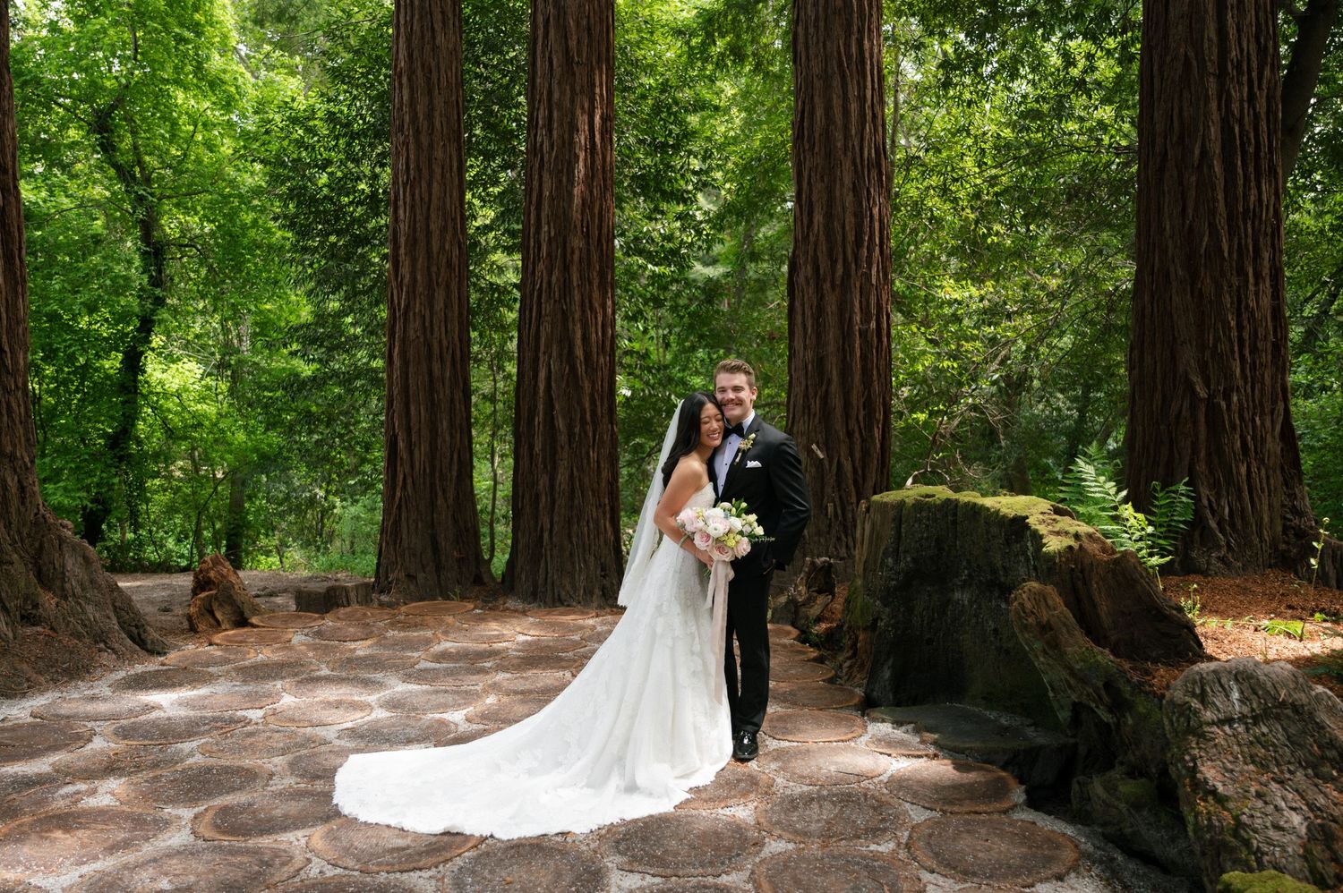 Bride and groom pose among towering redwood trees on a stone patio in a lush forest wedding setting.
