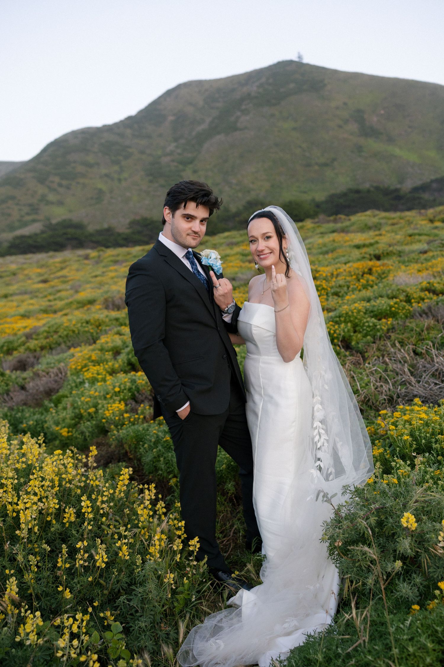Bride and groom posing in a wildflower field with green hills backdrop on their wedding day.