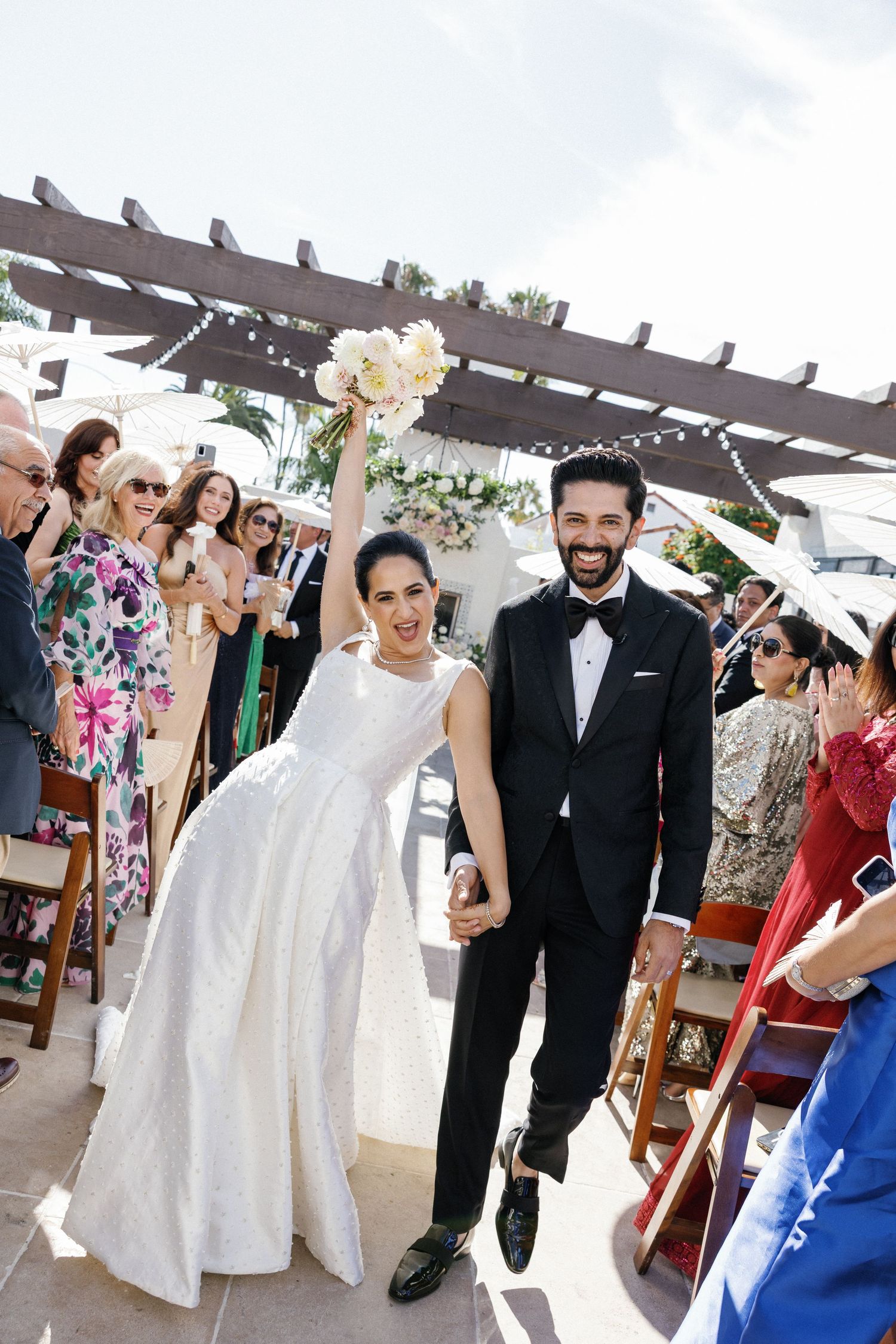 Wedding guests celebrate with newlyweds under a wooden pergola decorated with string lights during an outdoor reception.