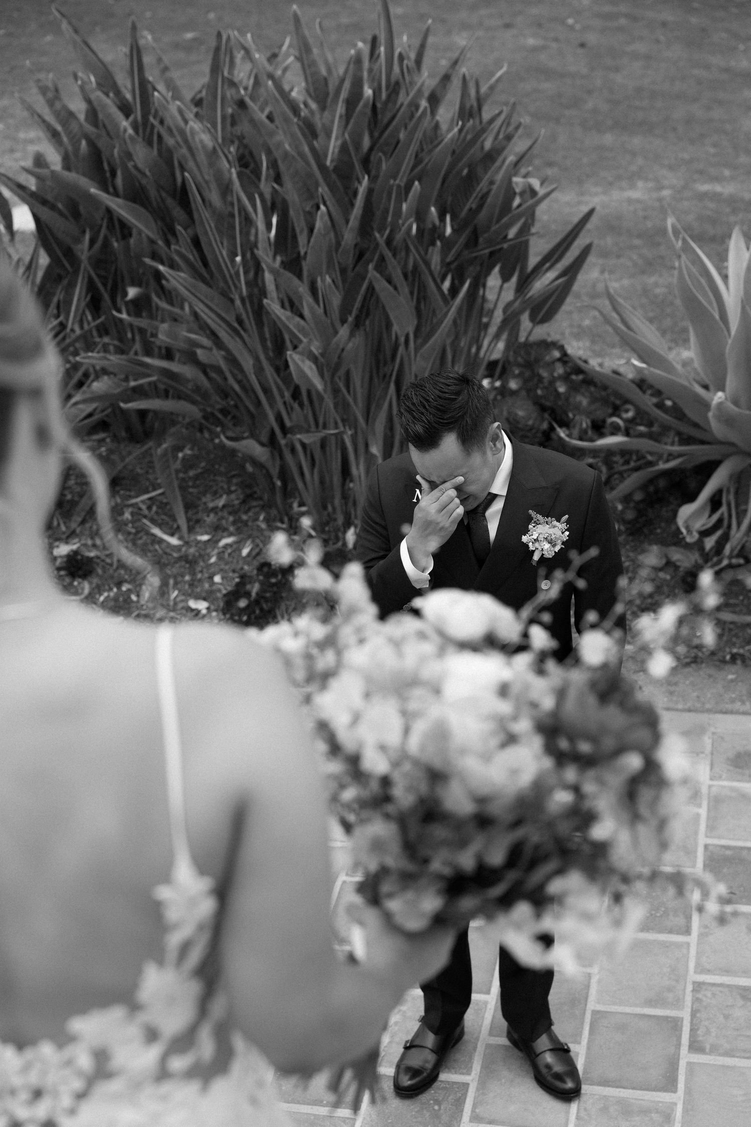 A black and white image shows dress shoes and formal attire against a stone walkway with plants in the background.