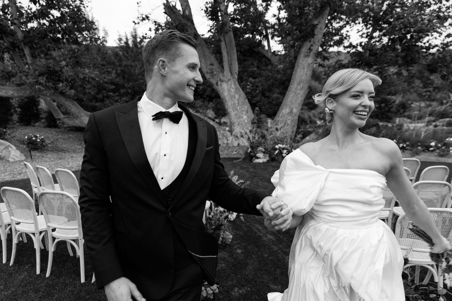 Black and white photo of a smiling bride and groom walking together outdoors at a garden wedding ceremony.