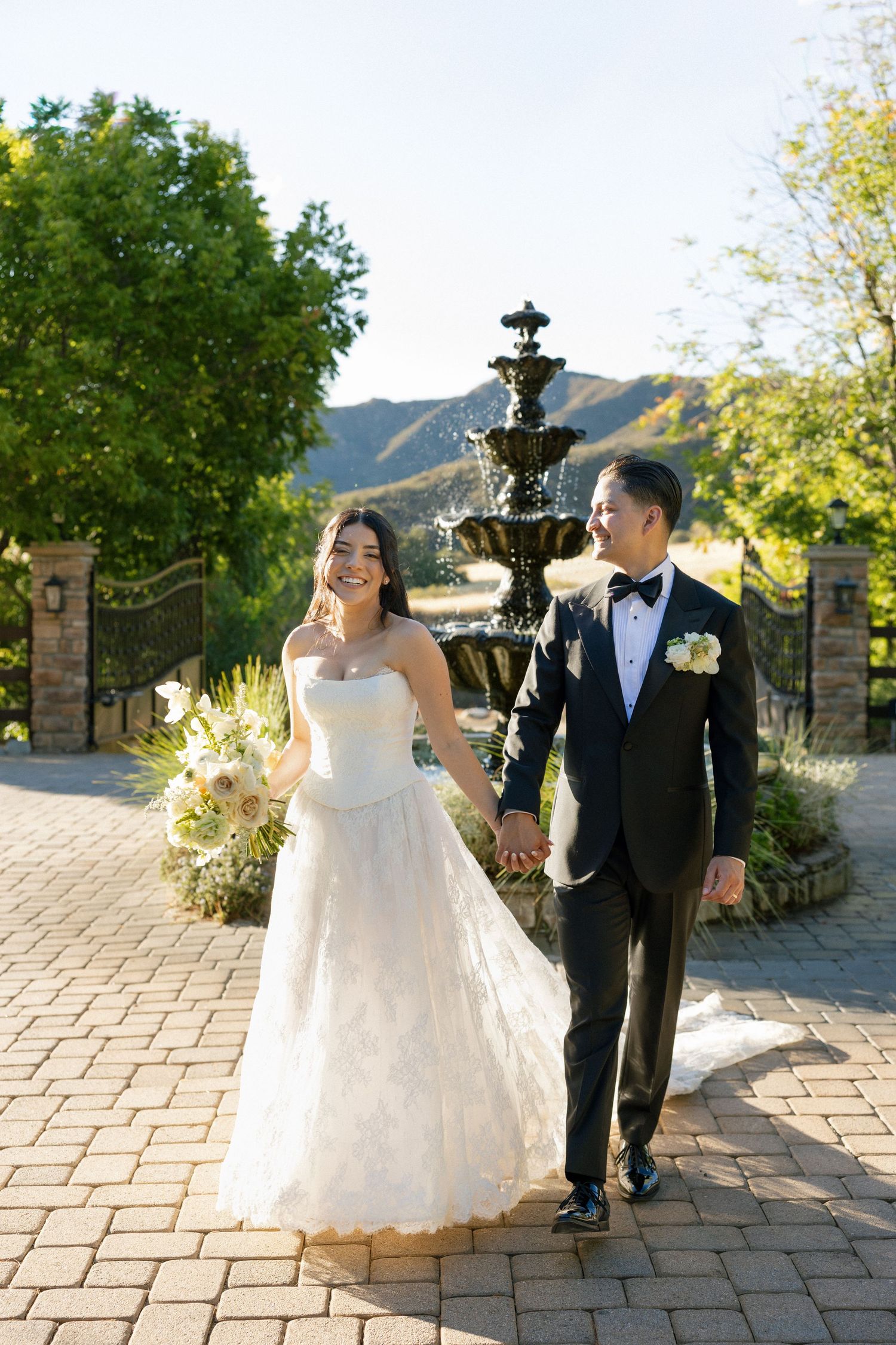 A couple in wedding attire walks along a stone path with a fountain and mountain backdrop during golden hour.