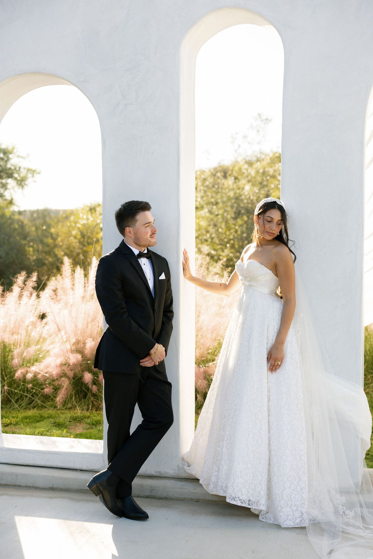 A wedding couple poses creatively in a white columned space with tall grass visible in the background at sunset.