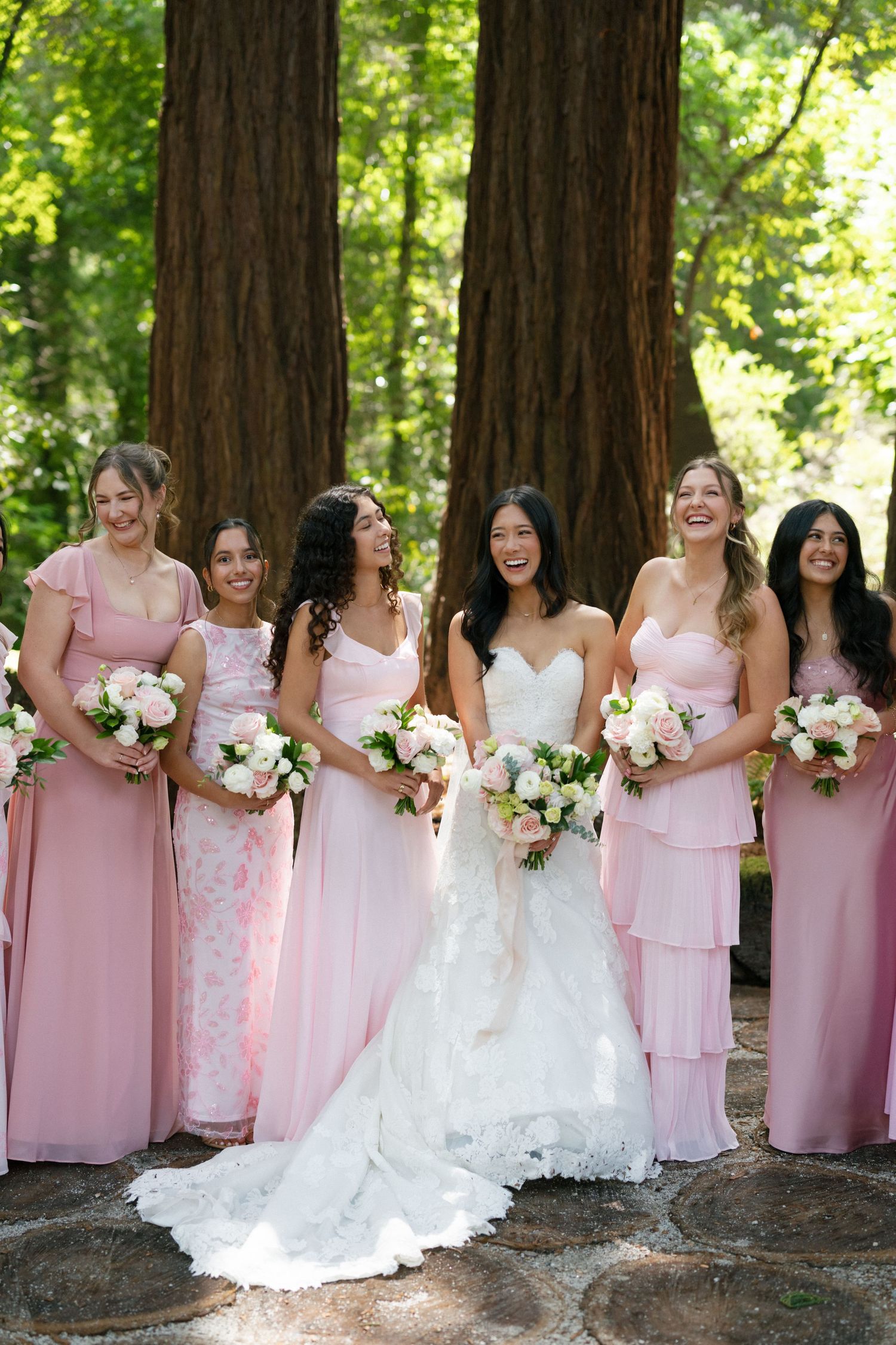Bride in white gown with bridesmaids in pink dresses holding bouquets, posing among redwood trees outdoors.