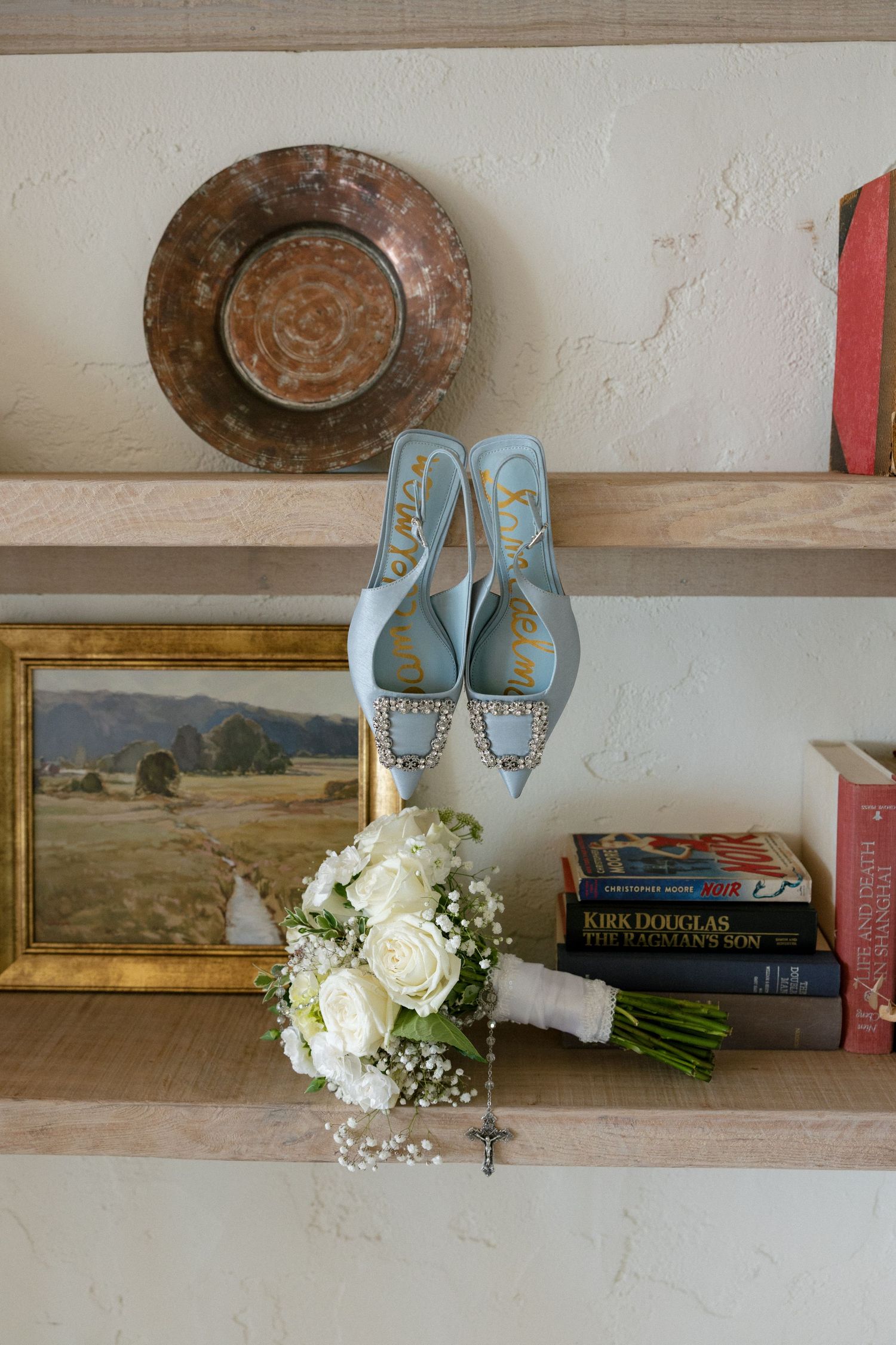 Light blue bridal heels displayed on wooden shelf with white rose bouquet, vintage painting, and stacked books at wedding venue.