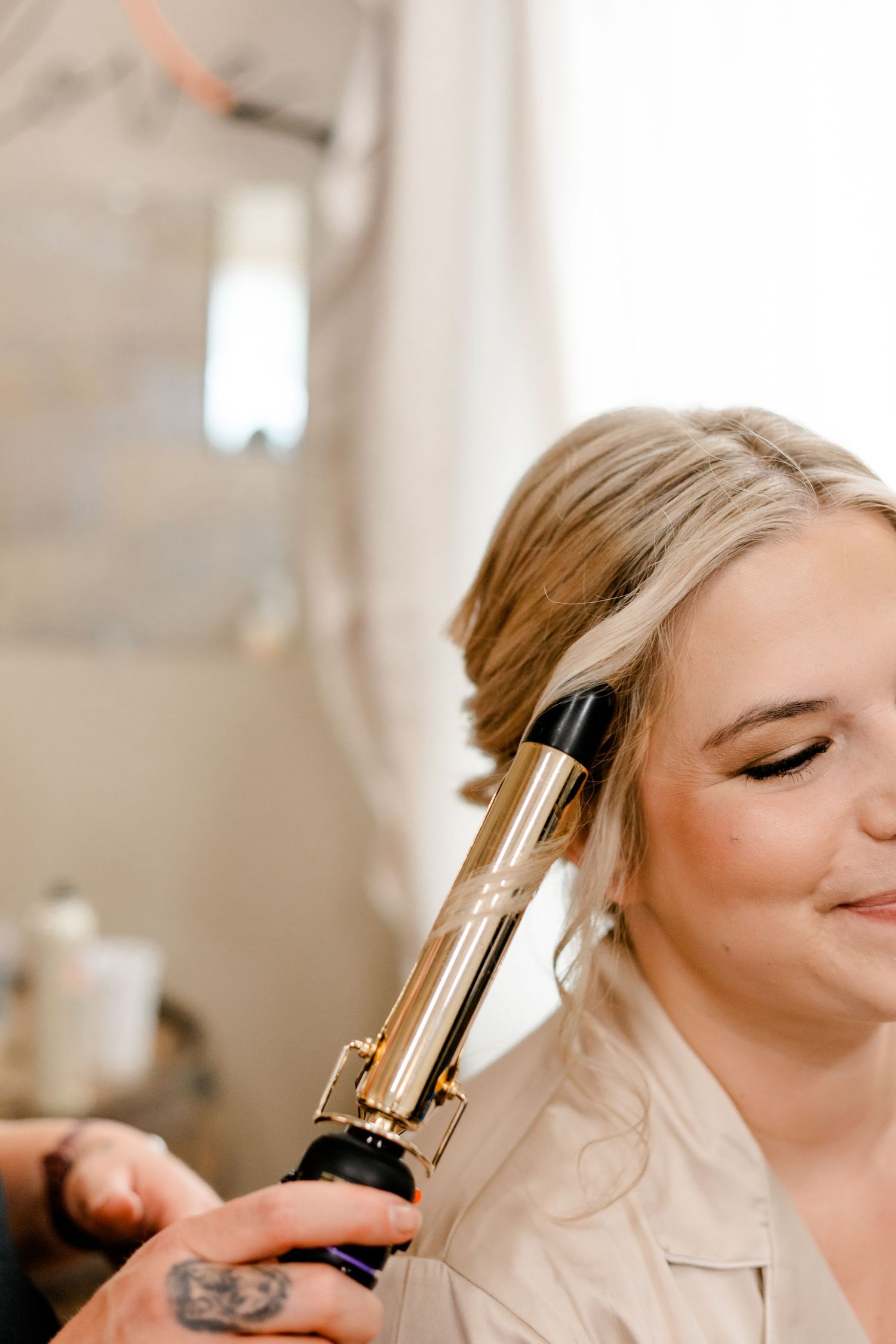 Blonde bride getting hair styled with gold curling iron during wedding day preparations.