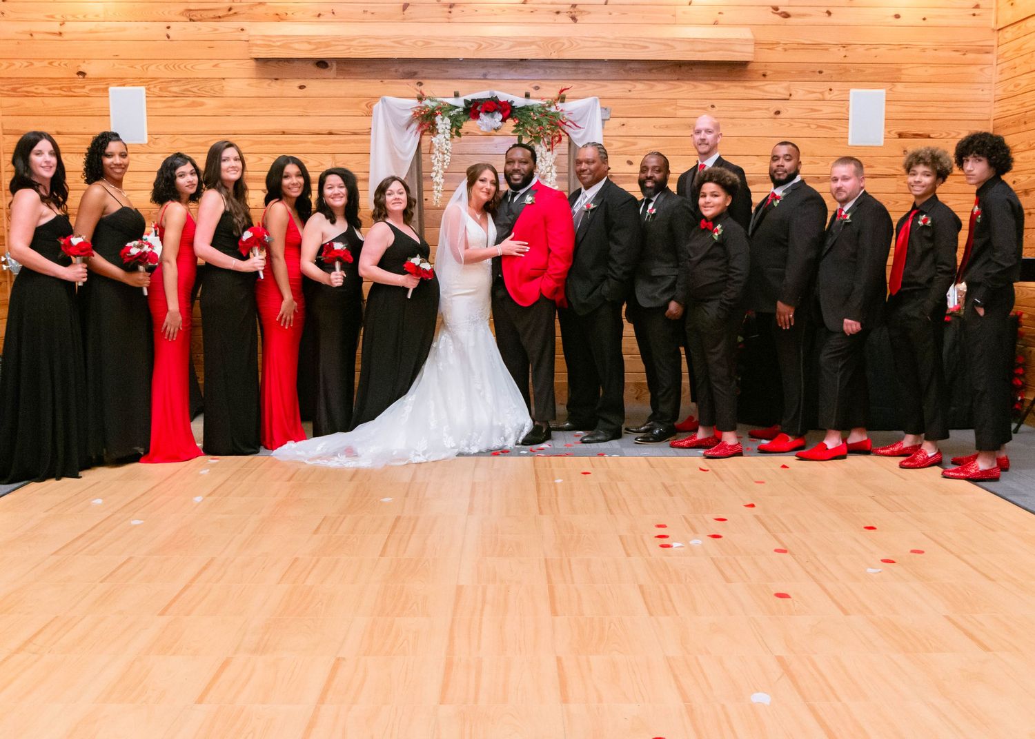 Large wedding party in red and black formal attire poses in line on wooden floor.