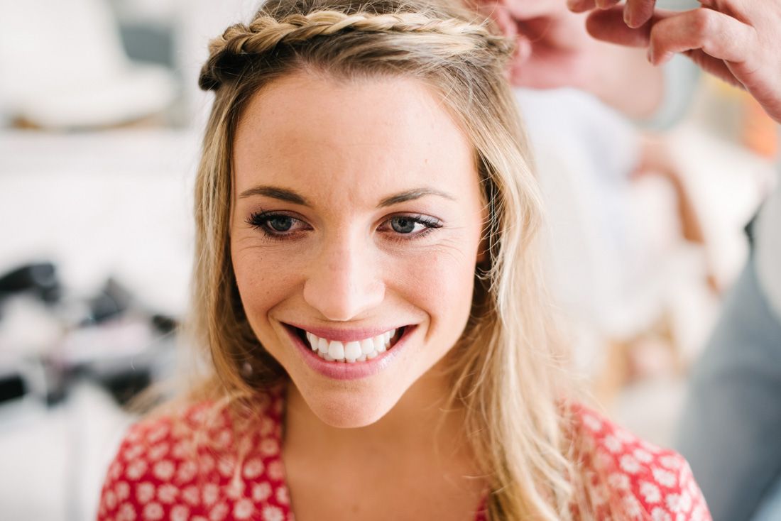 Smiling blonde woman with braided crown hairstyle being styled, wearing a red floral top.
