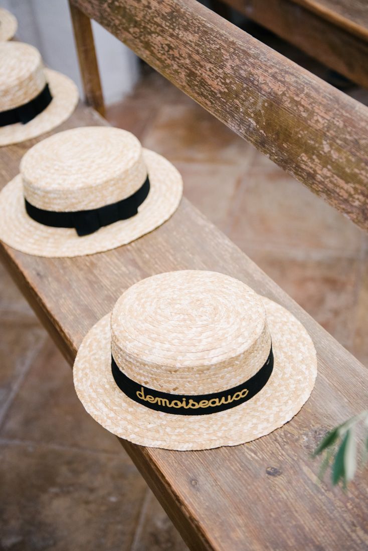 Straw boater hats with black ribbon bands reading demoiseau arranged on a wooden bench.
