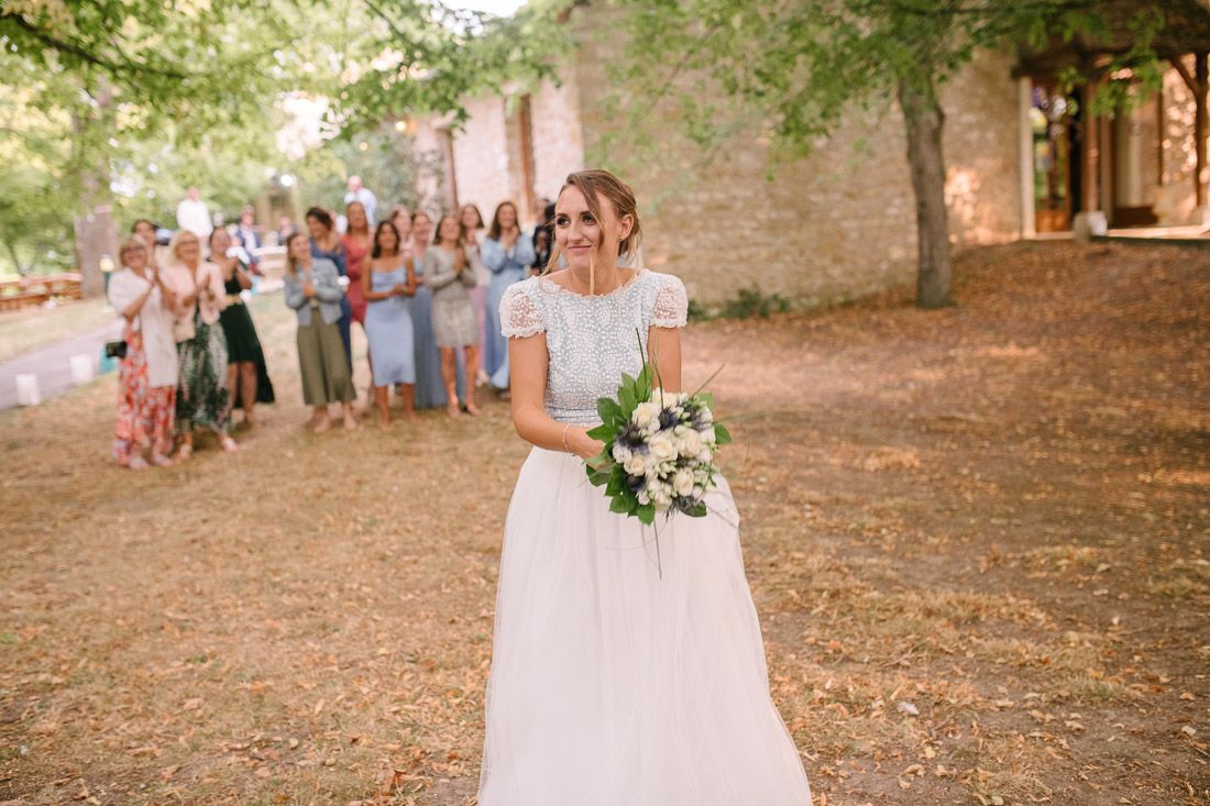 Smiling bride in lace wedding gown holds bouquet while guests applaud during outdoor bouquet toss ceremony.