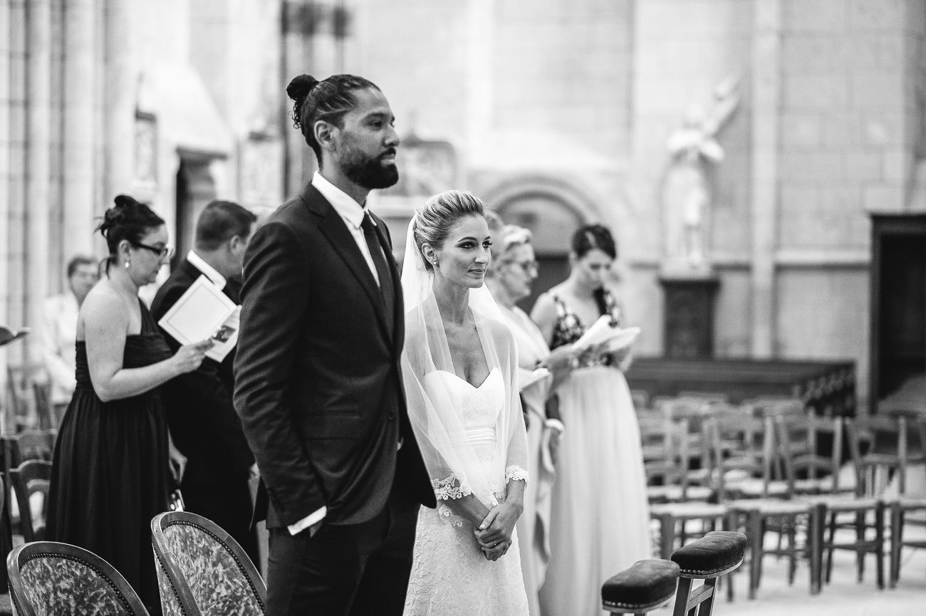 Black and white wedding ceremony photo of couple standing at church altar with guests seated in pews behind them.