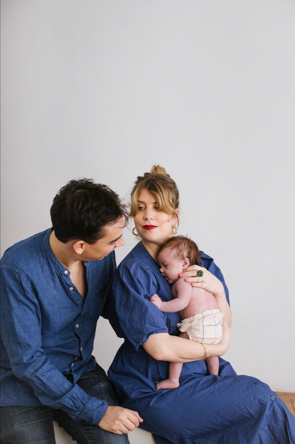 A couple dressed in blue sit together, tenderly looking at their sleeping newborn baby.