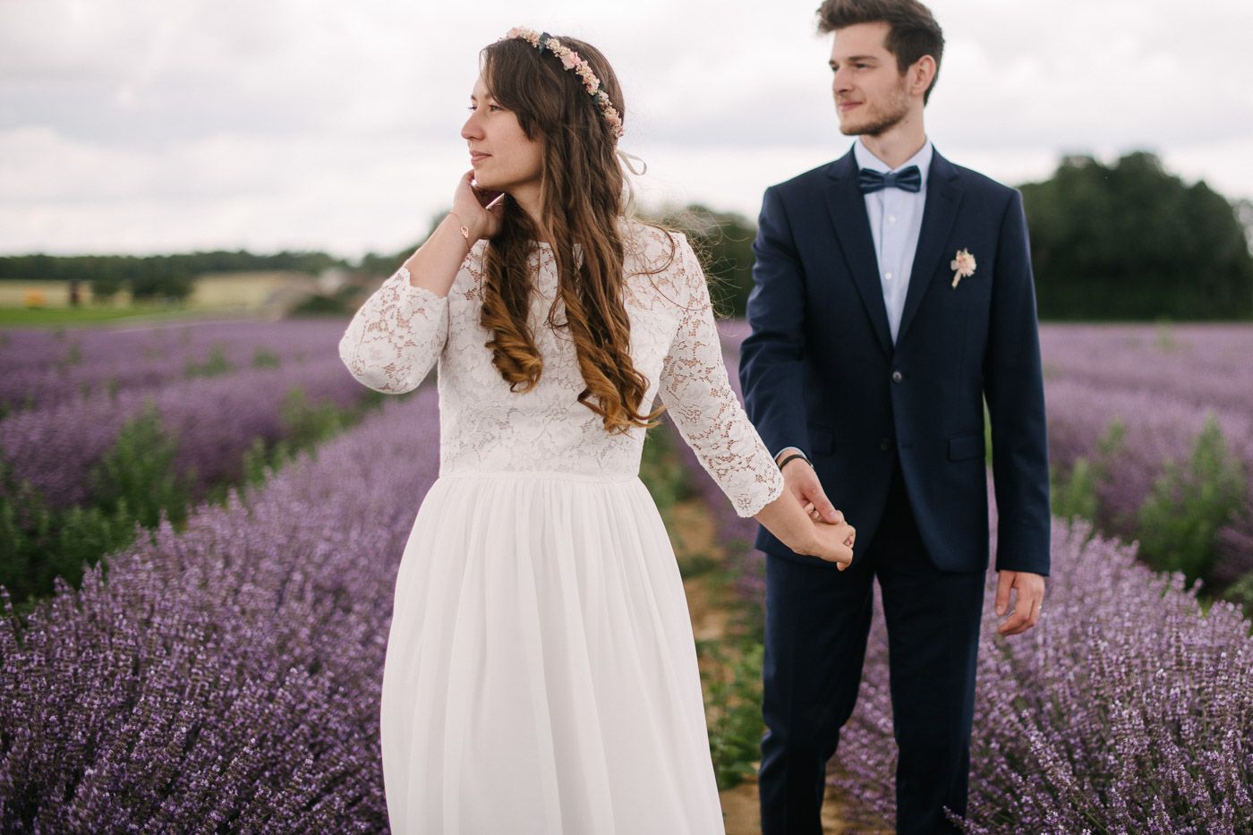 Bride and groom holding hands in a lavender field, bride in lace gown, groom in navy suit with bow tie.
