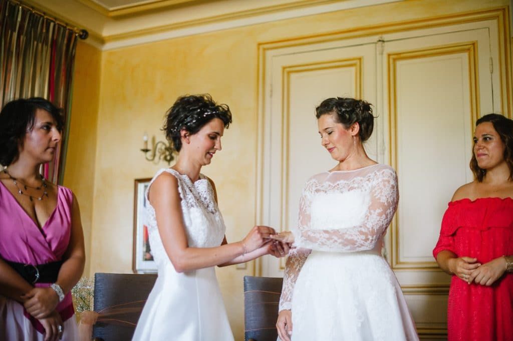 Two brides exchanging rings indoors with guests watching during ceremony.