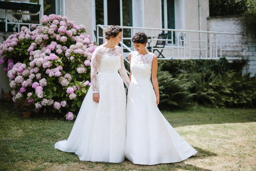 Two brides in white wedding gowns stand together in a garden with blooming pink hydrangeas in the background.