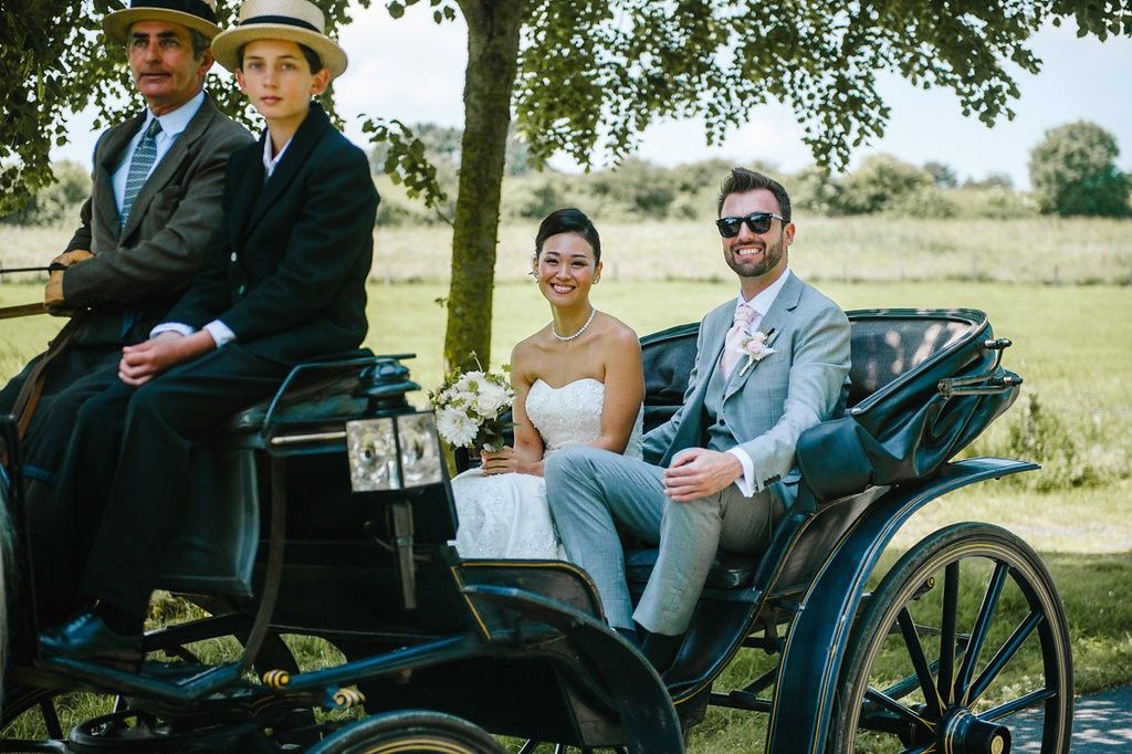 A bride and groom ride in a vintage black horse-drawn carriage with two coachmen on their wedding day.