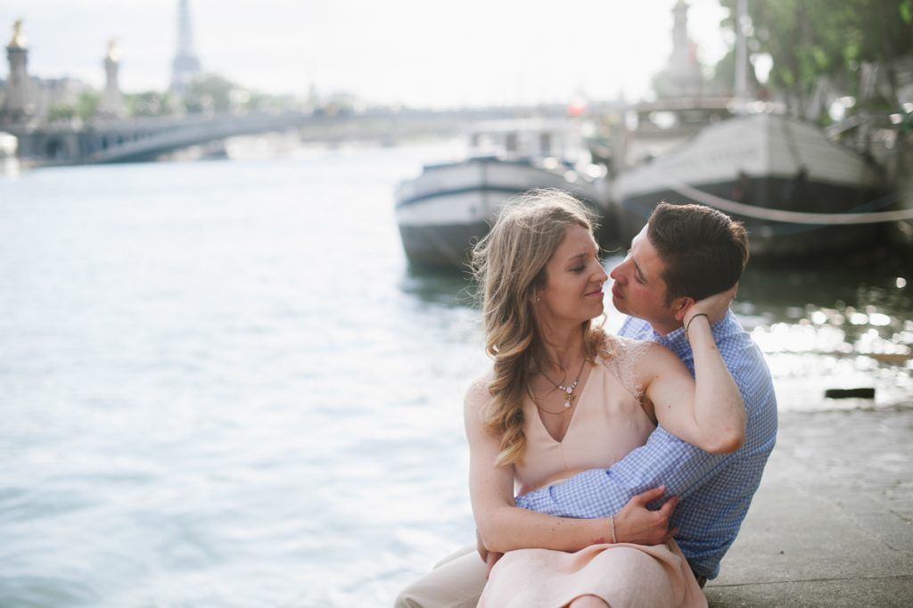 Couple embracing and sharing a kiss while seated by the Seine River with the Eiffel Tower behind.