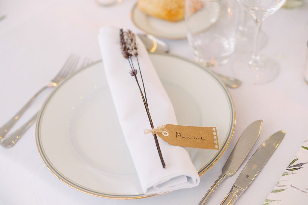 Close-up of a wedding place setting with white napkin, lavender sprig, and personalized name tag on gold-rimmed plate.