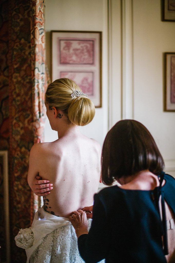 Assistant helping bride into lace wedding dress from behind near a draped window.