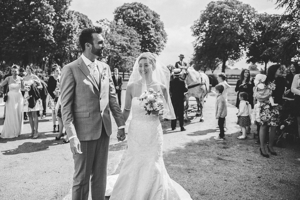 Bride and groom walk hand in hand down outdoor aisle in black and white wedding photo.