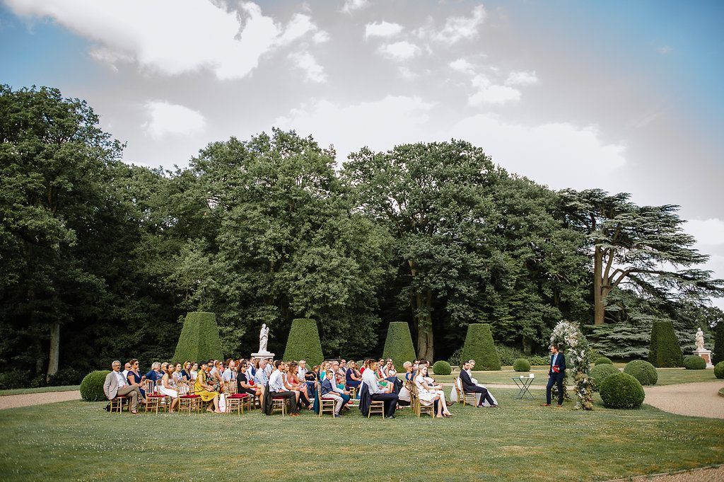 Wide aerial view of wedding guests seated for outdoor garden ceremony surrounded by tall topiary and lush trees.