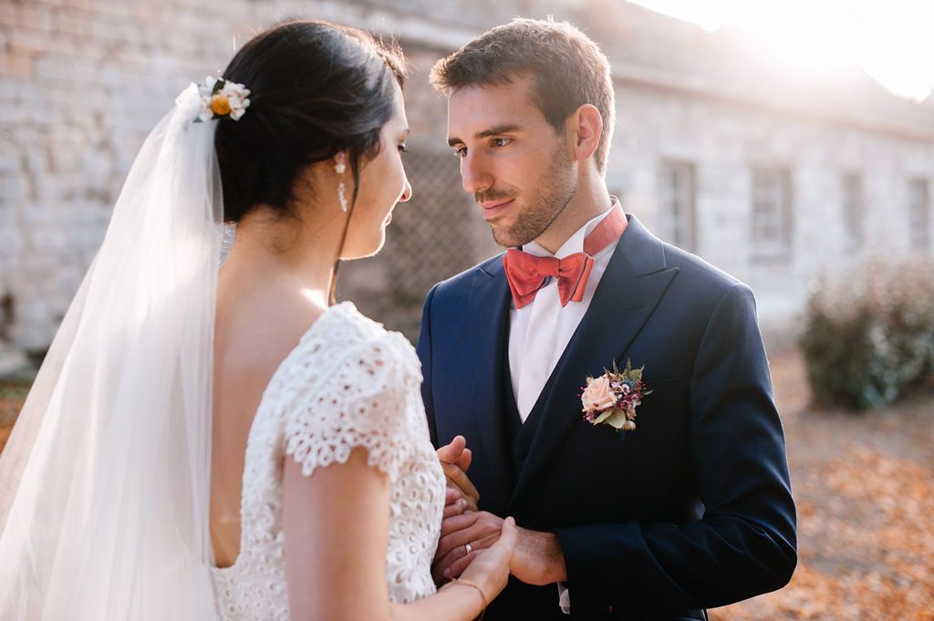 Groom in navy suit with red bow tie holds bride's hands during outdoor wedding portrait.