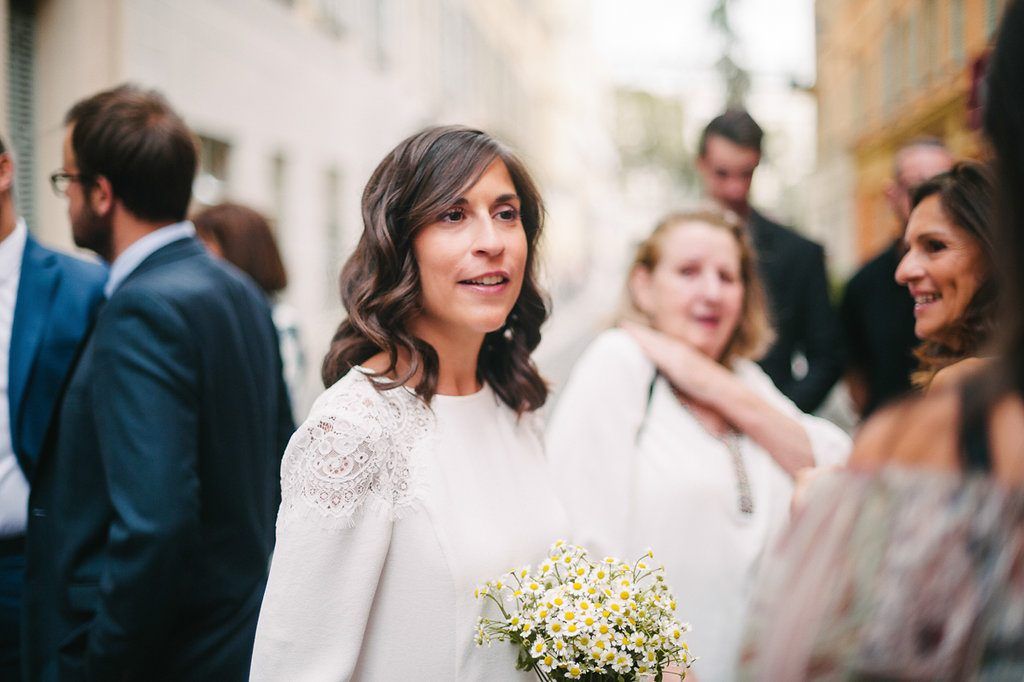 Radiant bride in white lace dress holds small bouquet among wedding guests outdoors.