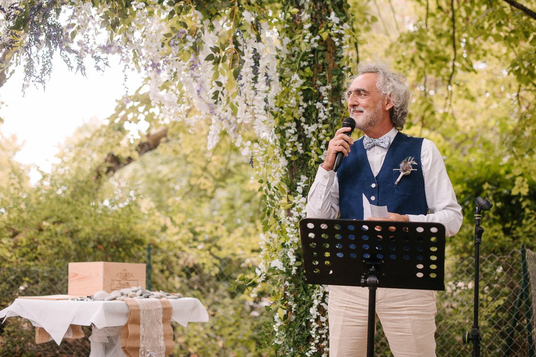 Elegant senior man speaking at outdoor wedding ceremony podium surrounded by lush greenery and white flowers.