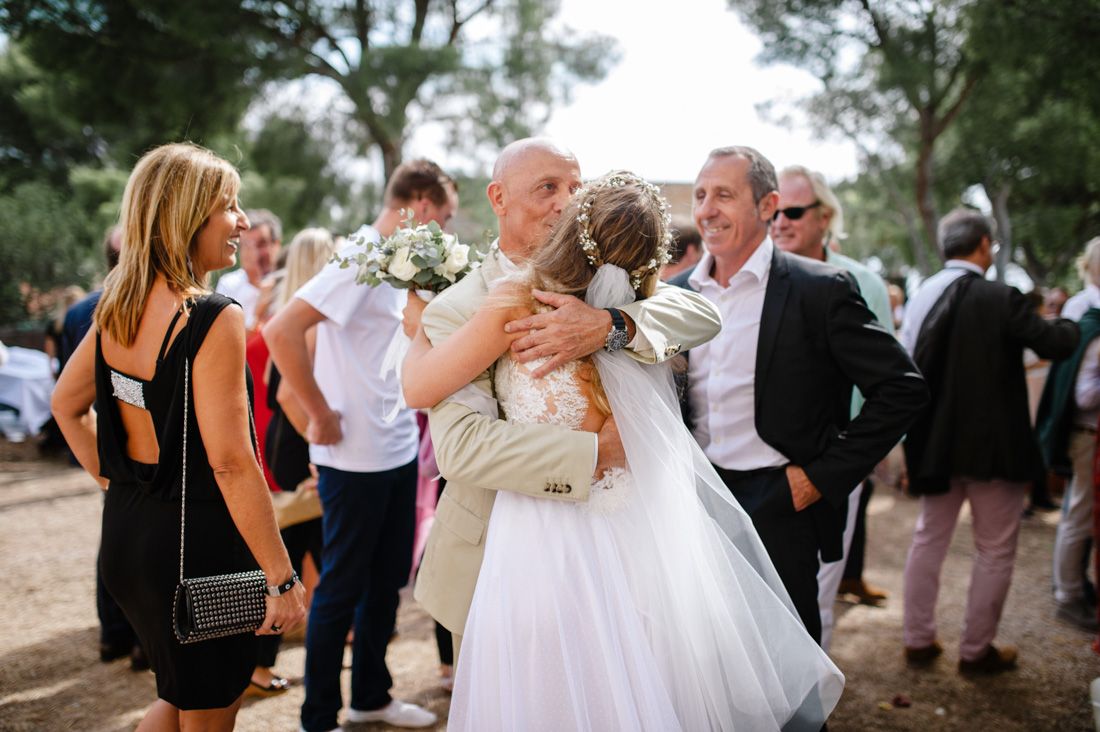 Bride in white dress hugging a bald man at an outdoor wedding reception surrounded by guests.