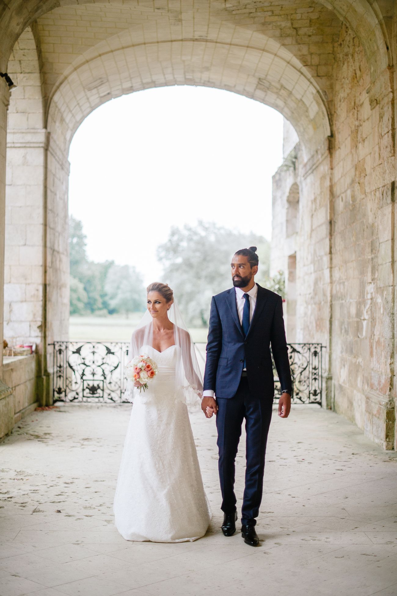 Bride and groom holding hands walking through a stone archway at a romantic outdoor wedding venue.