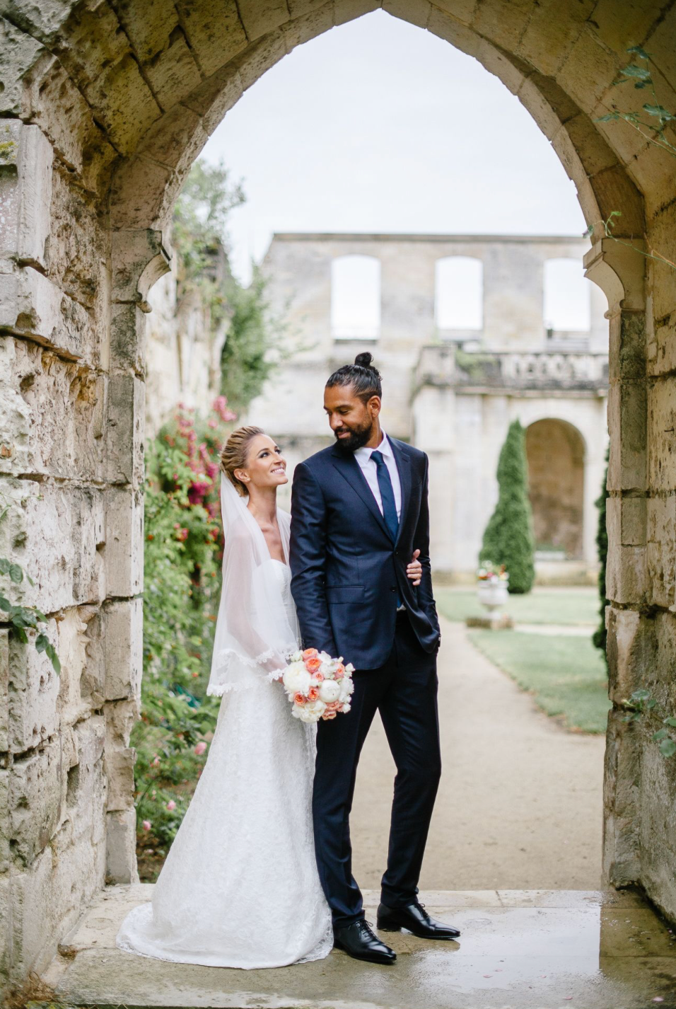 Elegant bride and groom posing under a stone archway at a historic venue, bride holding pink bouquet.