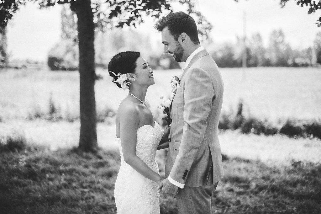 Black and white photo of a bride and groom holding hands and gazing at each other under a tree outdoors.