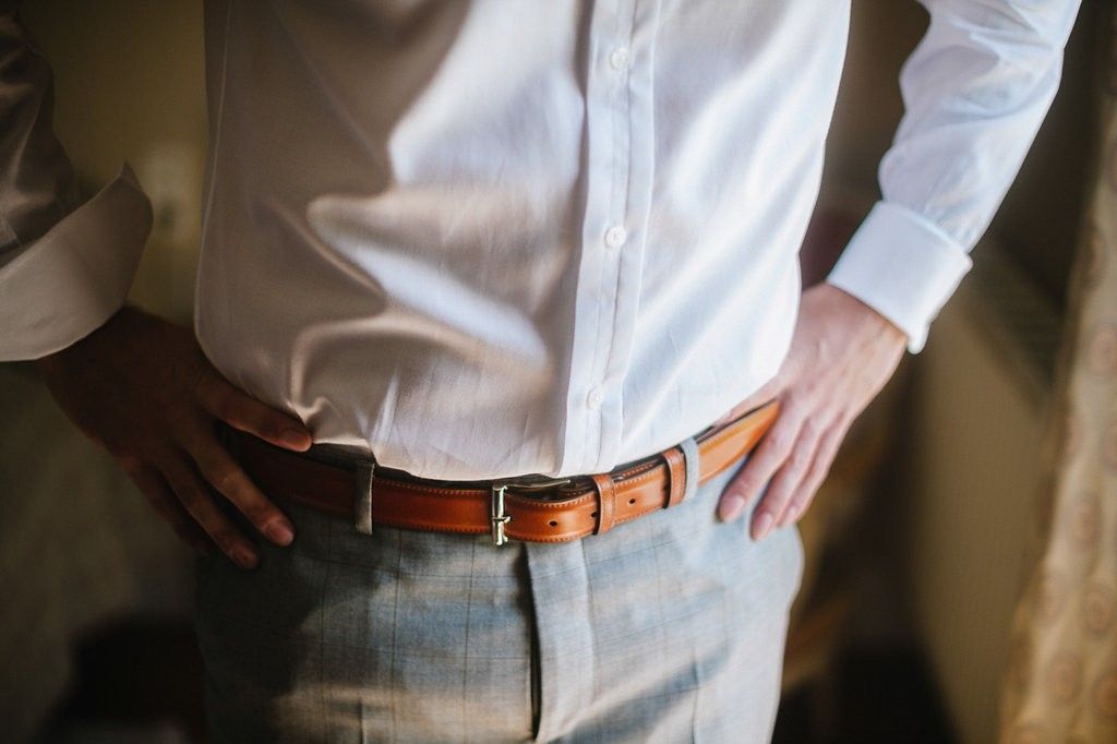 Groom in white shirt and tan leather belt with gray trousers on wedding day.