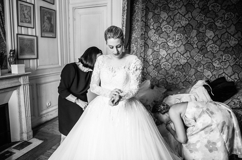 Bride in lace ball gown gets assistance from bridesmaid in elegant room during wedding preparations.