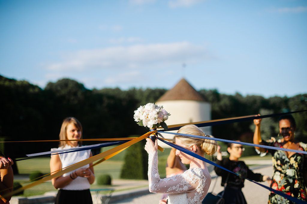 Bride tosses bouquet tied with colorful ribbons held by guests during outdoor wedding celebration.