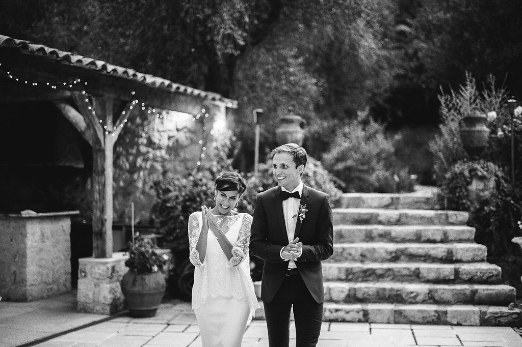 Black and white photo of bride and groom walking along stone terrace at dusk.