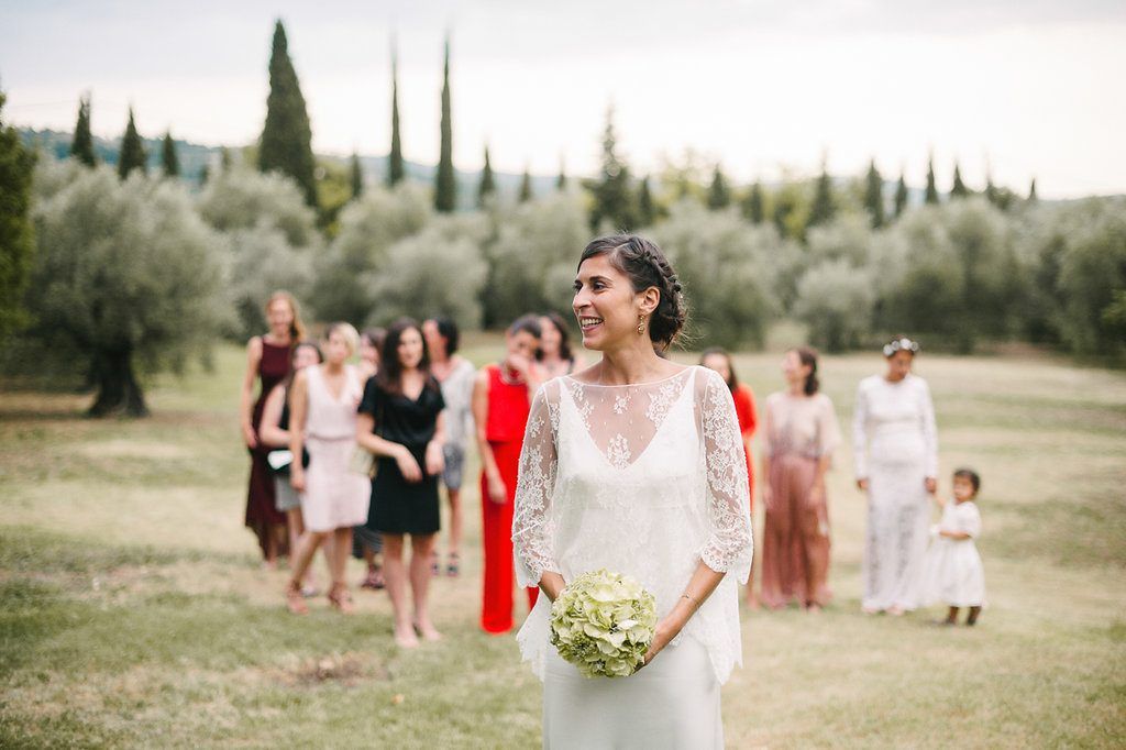 Smiling bride in lace wedding dress holding white bouquet walks outdoors with guests in background.