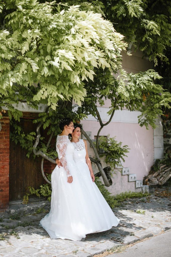 Two brides in white wedding gowns stand close together beneath a lush green tree on a cobblestone path.