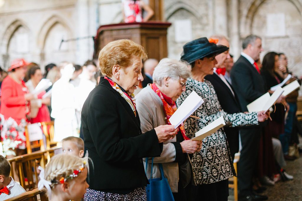 Wedding guests stand and read from programs during a church ceremony, dressed in elegant formal attire.