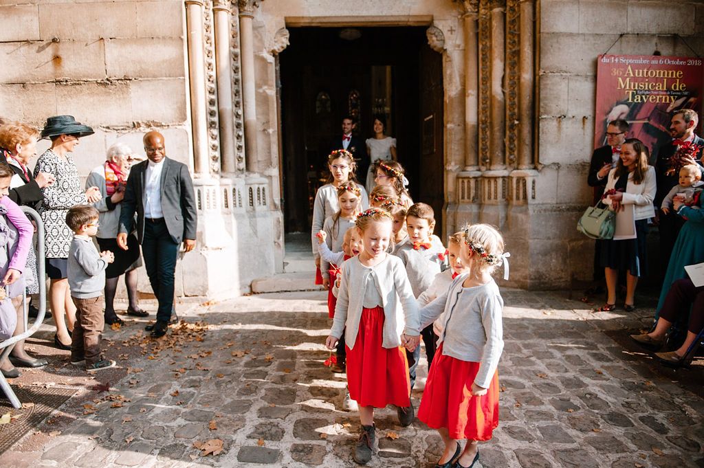 Flower girls in red and white outfits lead the wedding procession outside a historic stone church.