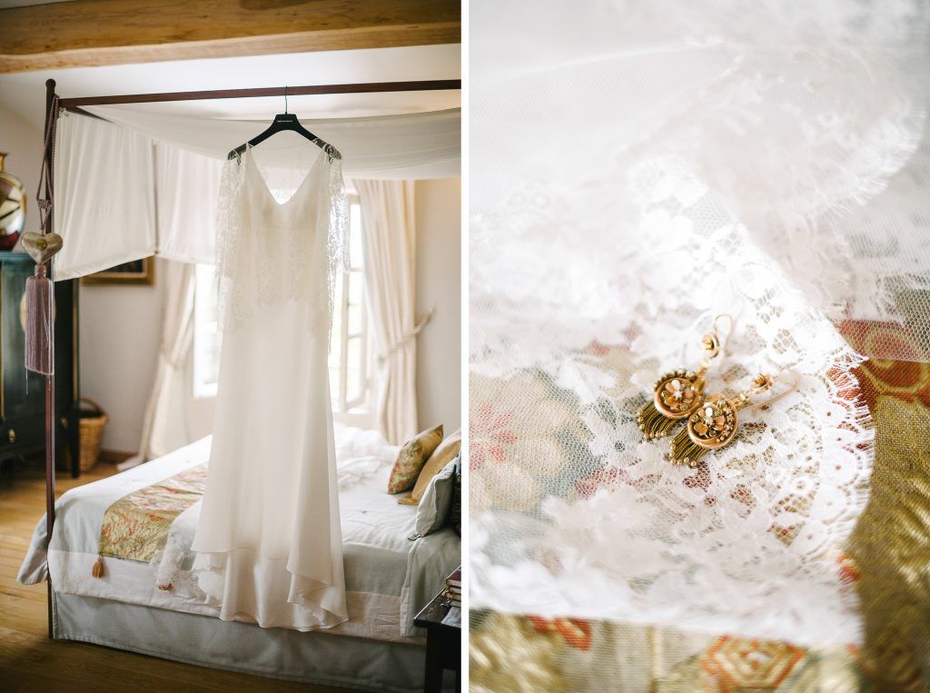 White wedding dress hanging on four-poster bed beside gold earrings on floral fabric.
