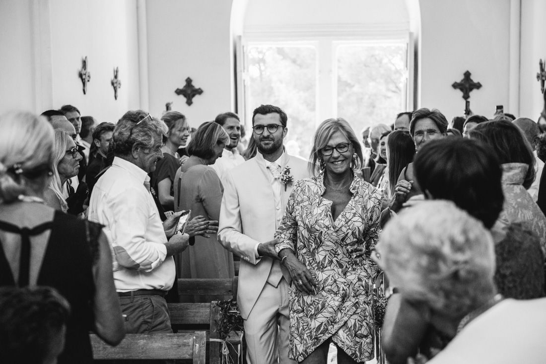Groom in beige suit walks down church aisle with smiling guest at a crowded wedding ceremony.