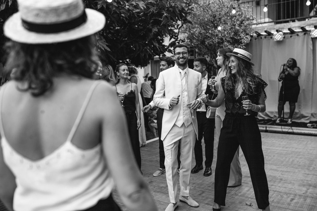 Groom in white suit socializing with wedding guests outdoors in black and white photo.